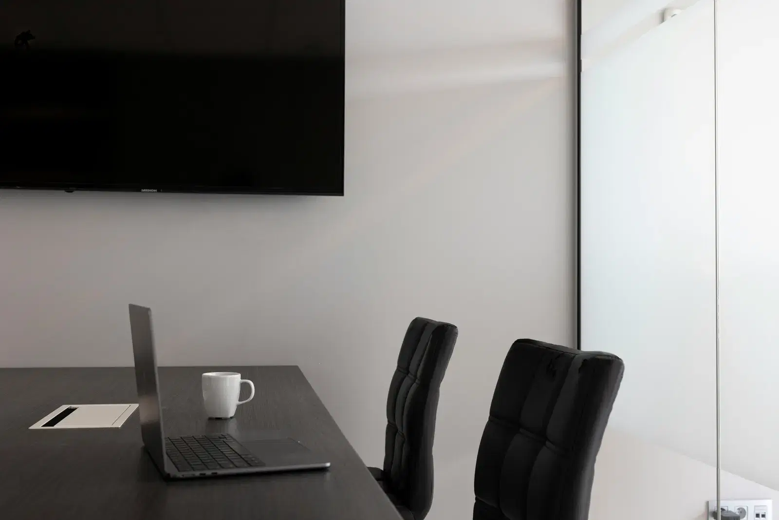 A sleek, modern office setup with a laptop, cup, and TV in an empty conference room.